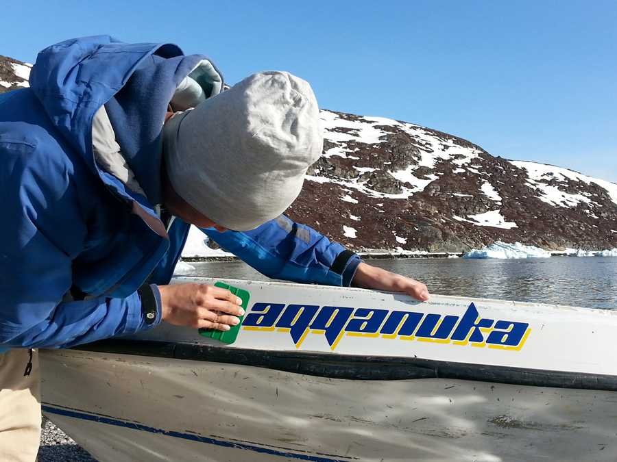Her ses båden aqqanuka i Grønland under en smuk blå himmel. Bådens navn er lavet i en mørkeblå folie og har skygge i gul.